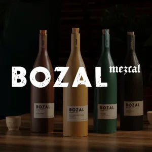 Vier Bozal‑Mezcal‑Flaschen in verschiedenen Farben (dunkelbraun, gelb, grün und schwarz) stehen nebeneinander auf einer Holzoberfläche vor einem dunklen Hintergrund.