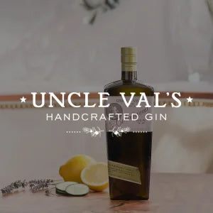 „Eine dunkelflasche mit der Aufschrift ‚Uncle Val’s Handcrafted Gin‘ steht auf einem hellen Holztisch; daneben liegen Zitronenhälften und Kräuterzweige vor einem neutralen, weich beleuchteten Hintergrund.